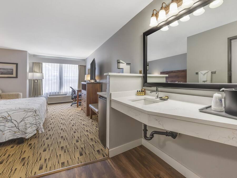 A hotel room with a large bed, a sitting area with a chair and a table, a large mirror, and a bathroom with a sink and a shower.