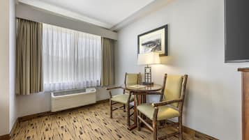 Een hotelkamer met een groot raam, een ronde tafel met twee stoelen, een wandlamp en een lijst met een afbeelding.