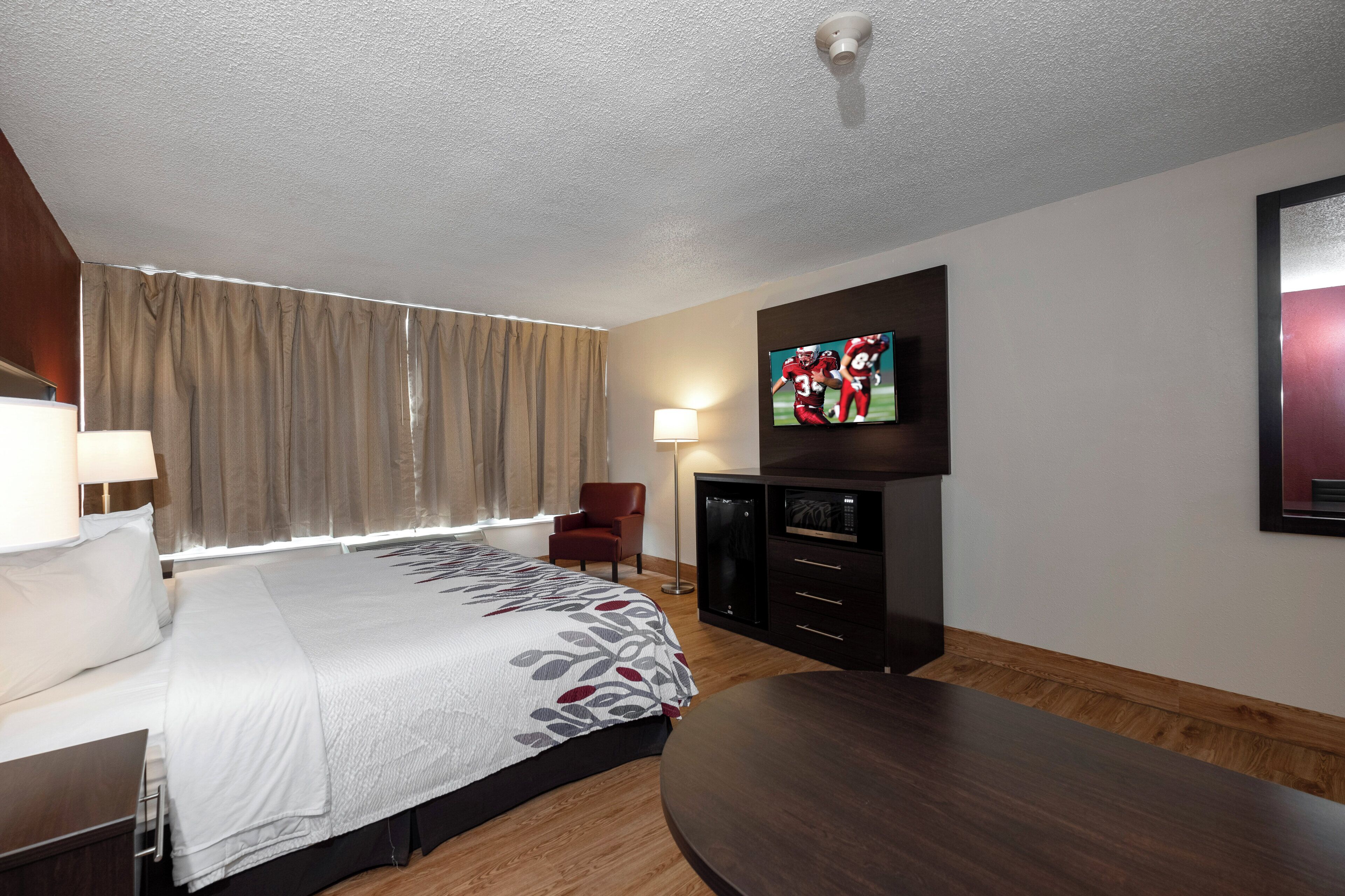 Une chambre d’hôtel avec un lit, un téléviseur fixé au mur, un bureau, une chaise et un plancher en bois.