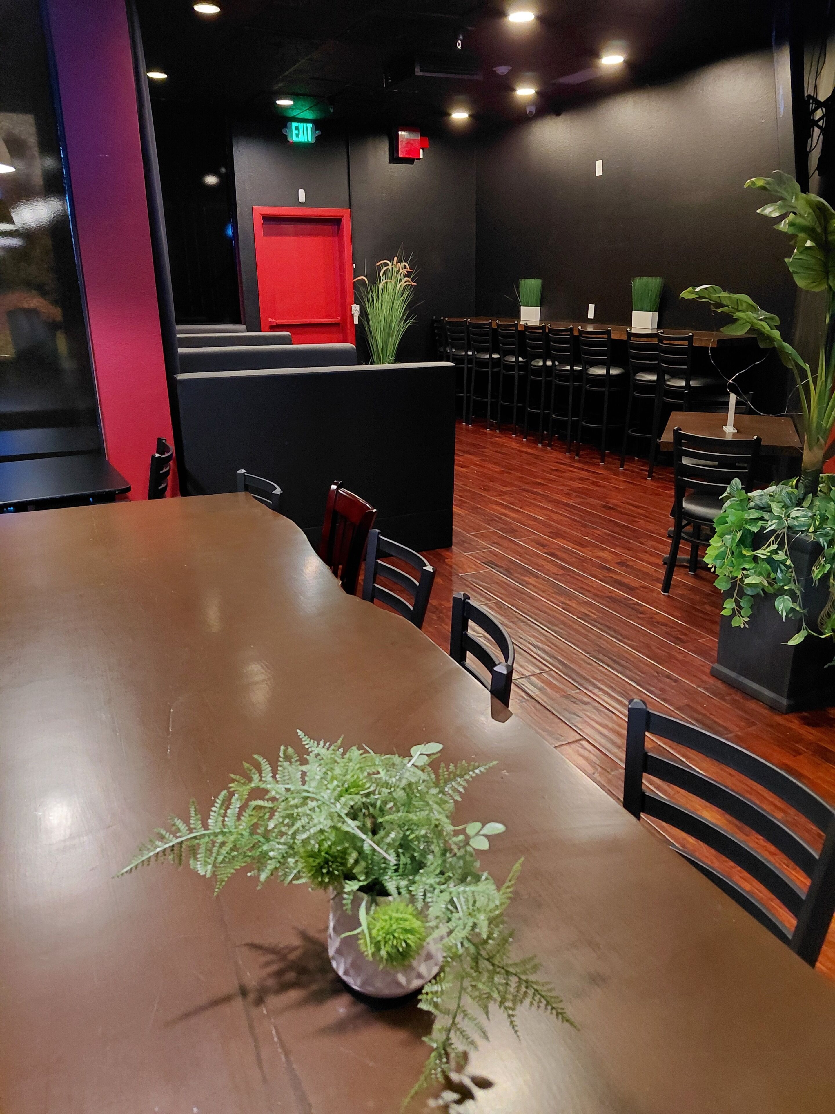 L’intérieur d’un restaurant avec une longue table en bois, des chaises, des plantes en pot et un panneau de signalisation rouge indiquant la sortie, fixé au mur.