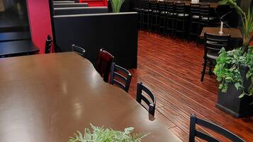 A restaurant interior with a long wooden table, chairs, potted plants, and a red exit sign on the wall.