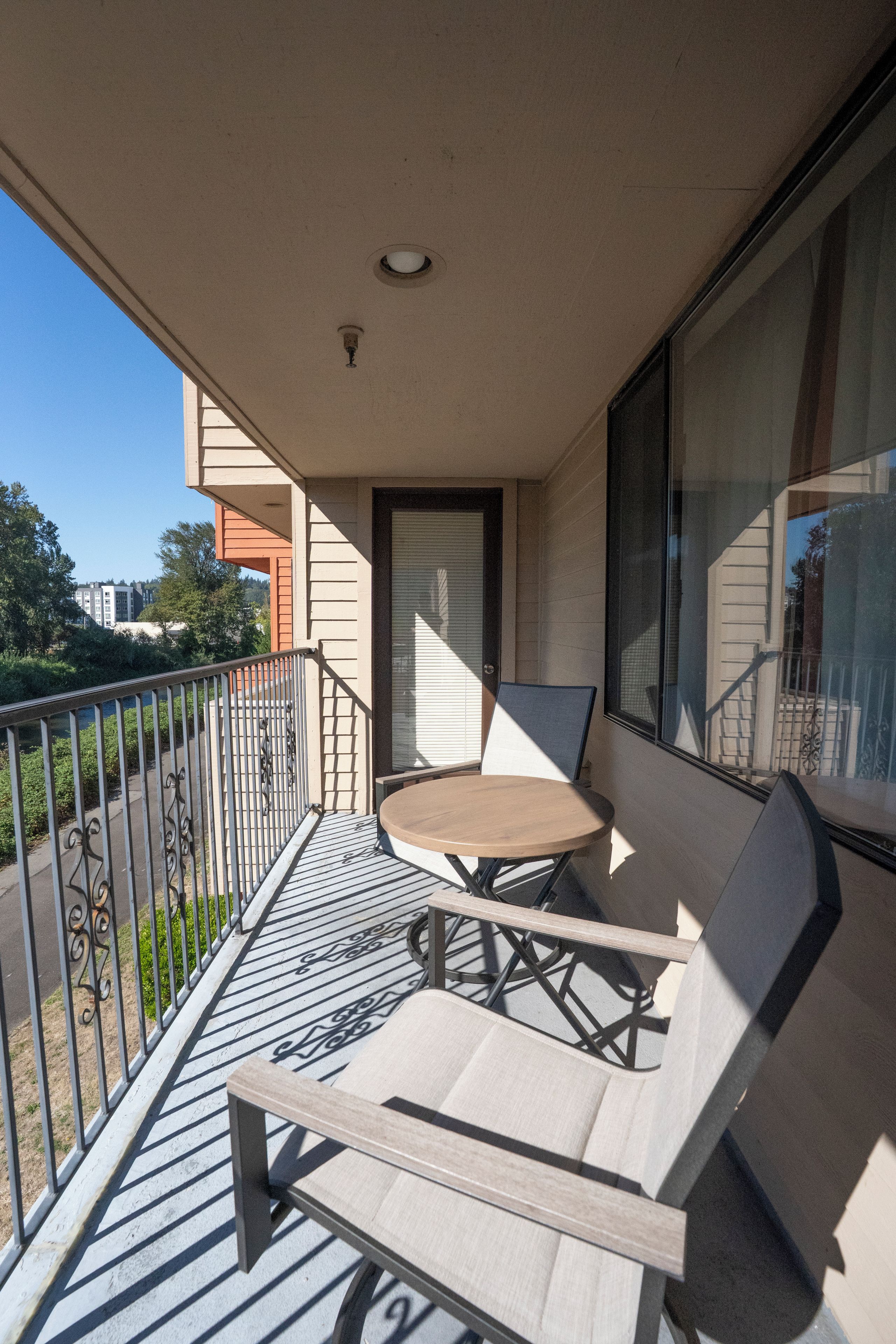 Een balkon met een tafel en twee stoelen, een raam met zonwering, en een metalen hekwerk.