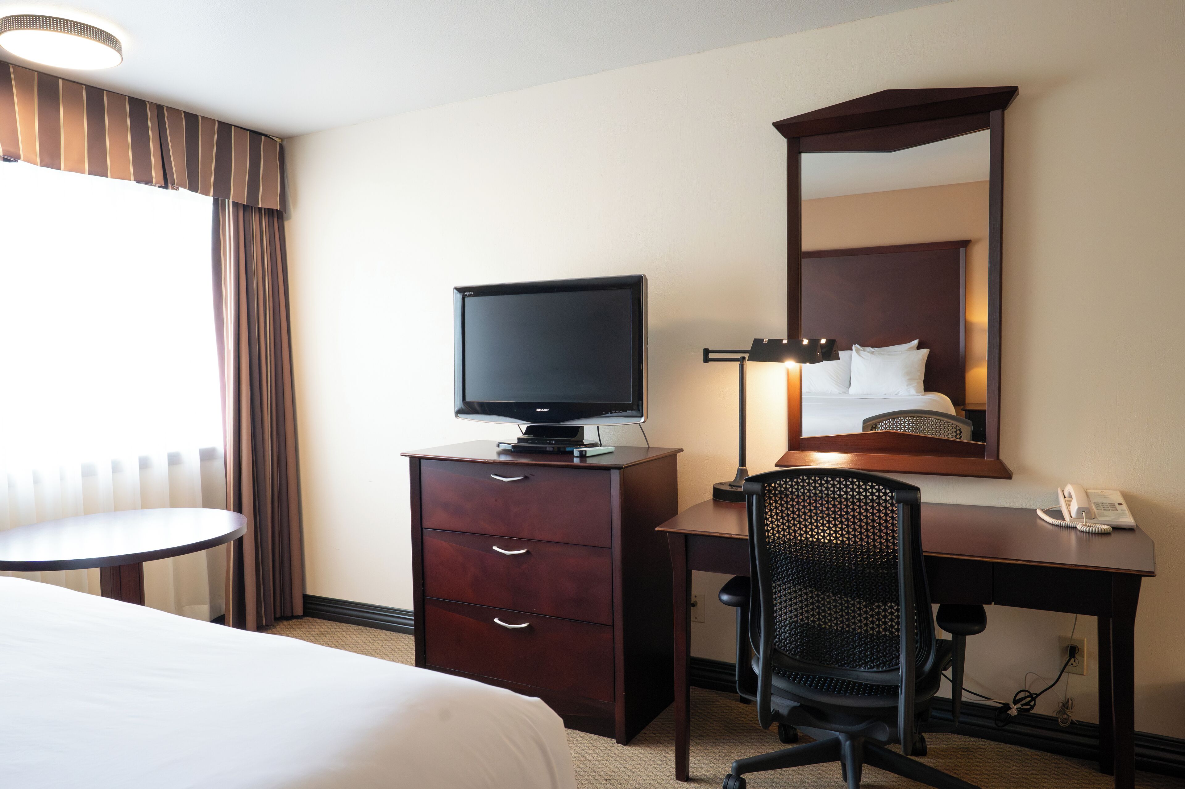 A hotel room with a bed, a desk with a computer, a chair, a television on a dresser, a mirror, and a telephone.