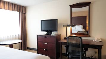 A hotel room with a bed, a desk with a computer, a chair, a television on a dresser, a mirror, and a telephone.