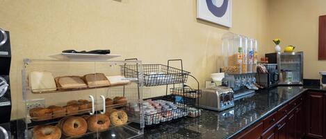 A breakfast bar with bread, muffins, and bagels. There are also coffee makers and a fruit display.