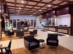 A hotel lobby with a reception desk, seating area, and a kitchen in the background.