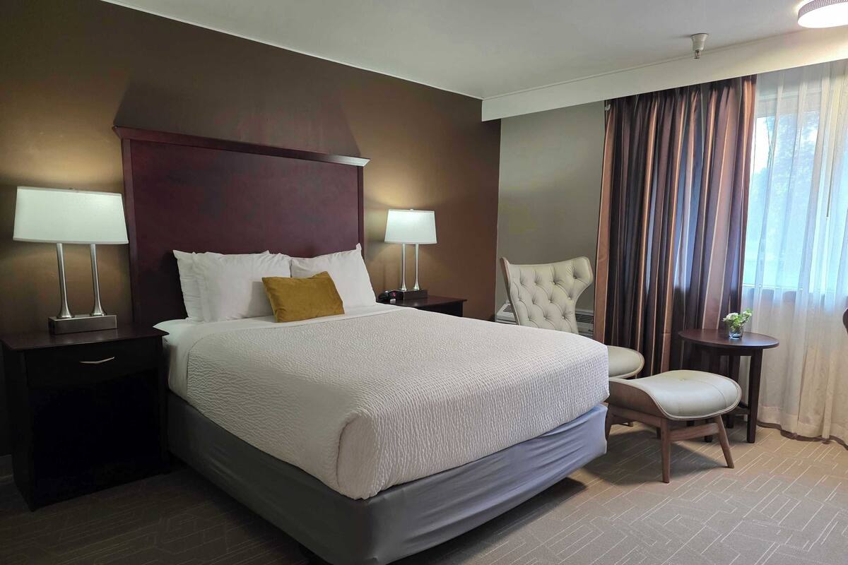 Hotelkamer met een groot bed, twee bedlampen, een gestoffeerde fauteuil, een tafeltje met een vaas, en uitzicht op het buitenlandschap door gordijnen.