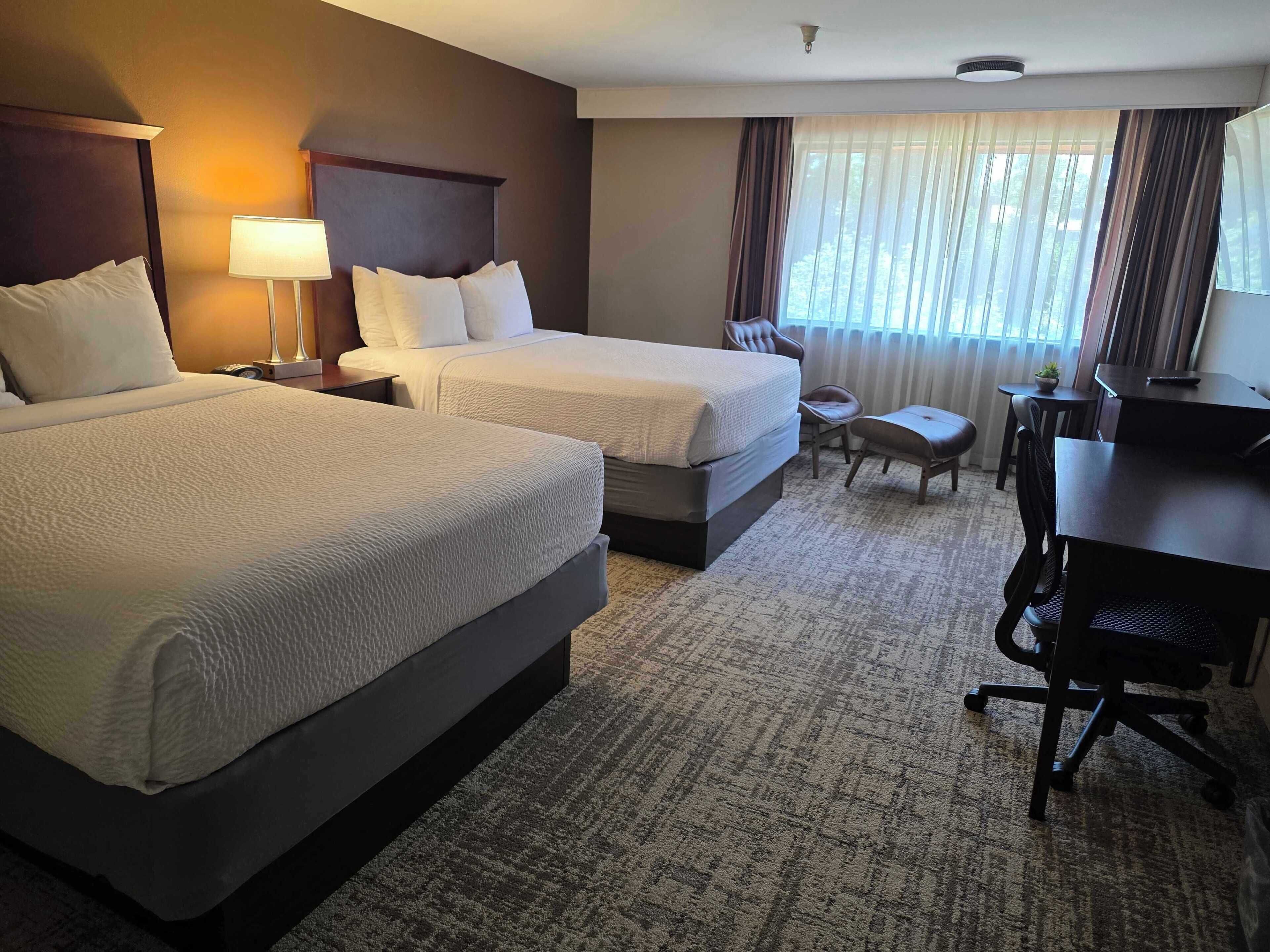 A hotel room with two beds, a desk, a chair, a lamp, and a view of the outdoors.