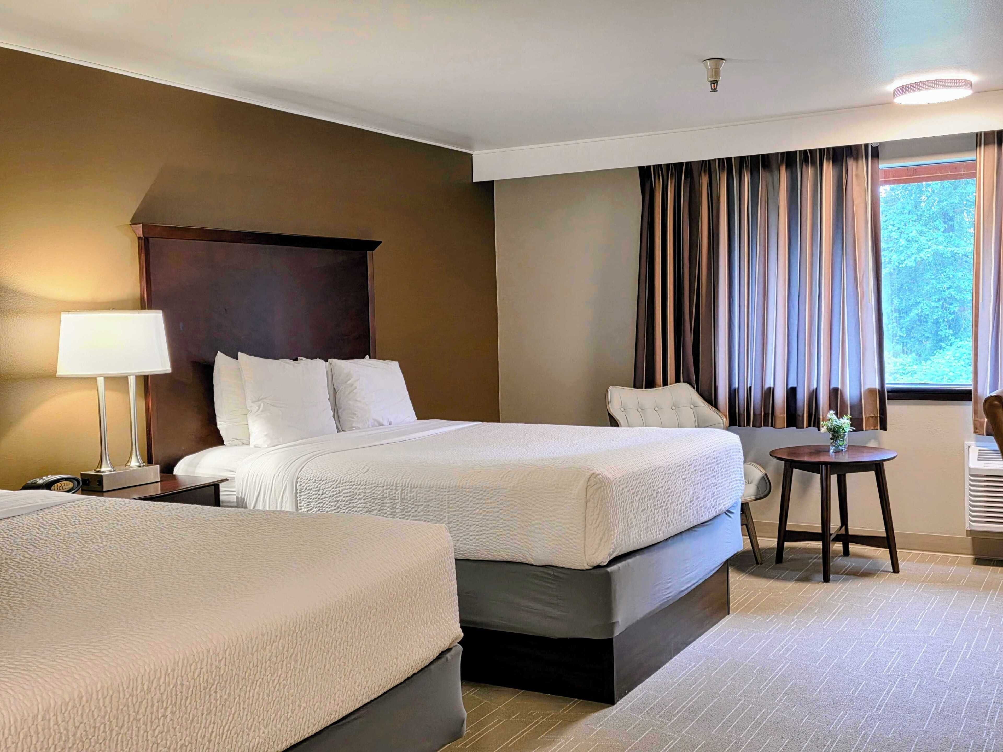A hotel room with two beds, a bedside table with a lamp, a small round table with a vase, and a window with curtains.