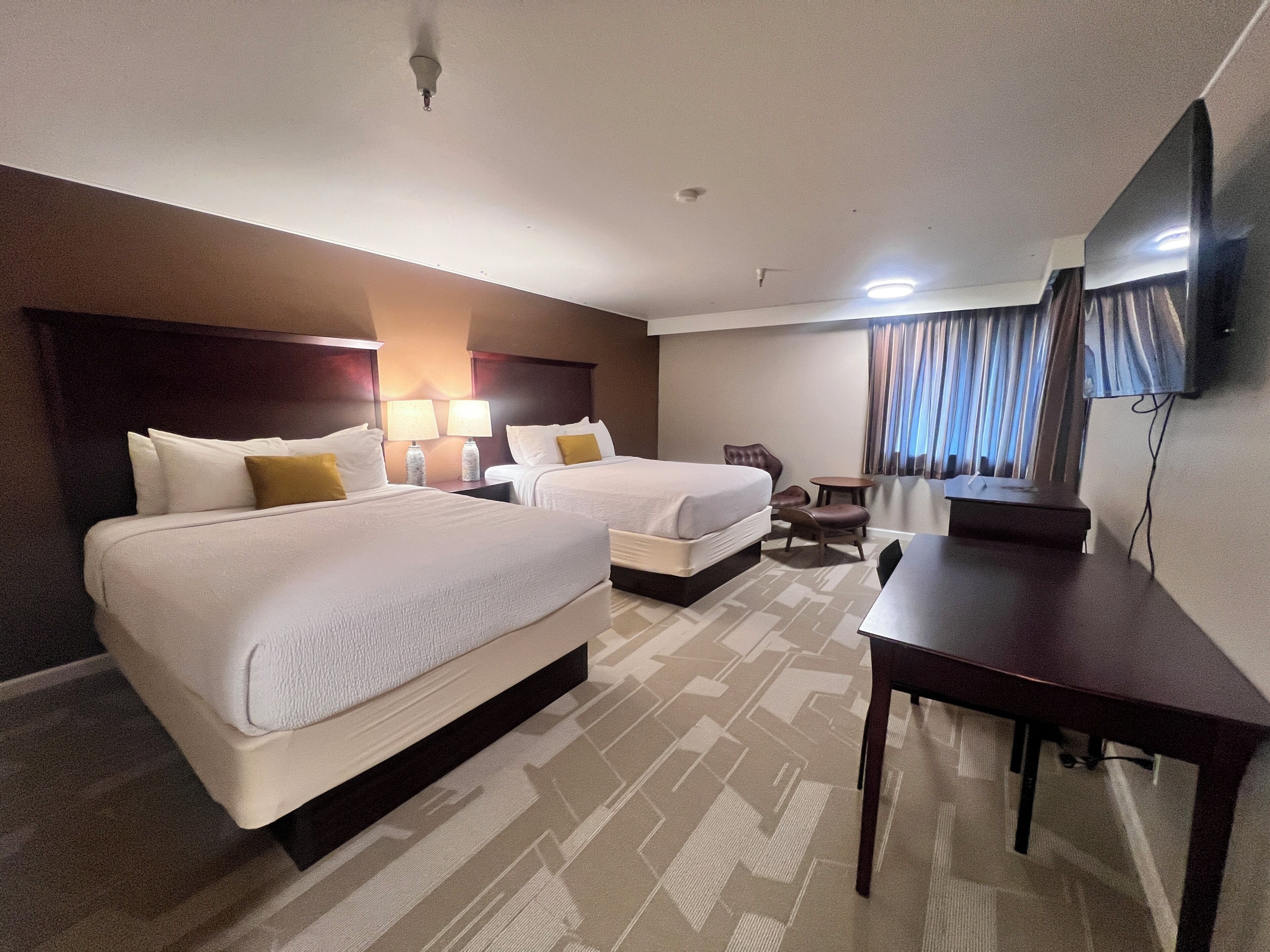 A hotel room with two beds, a desk, a chair, a lamp, a TV, and a window with curtains.
