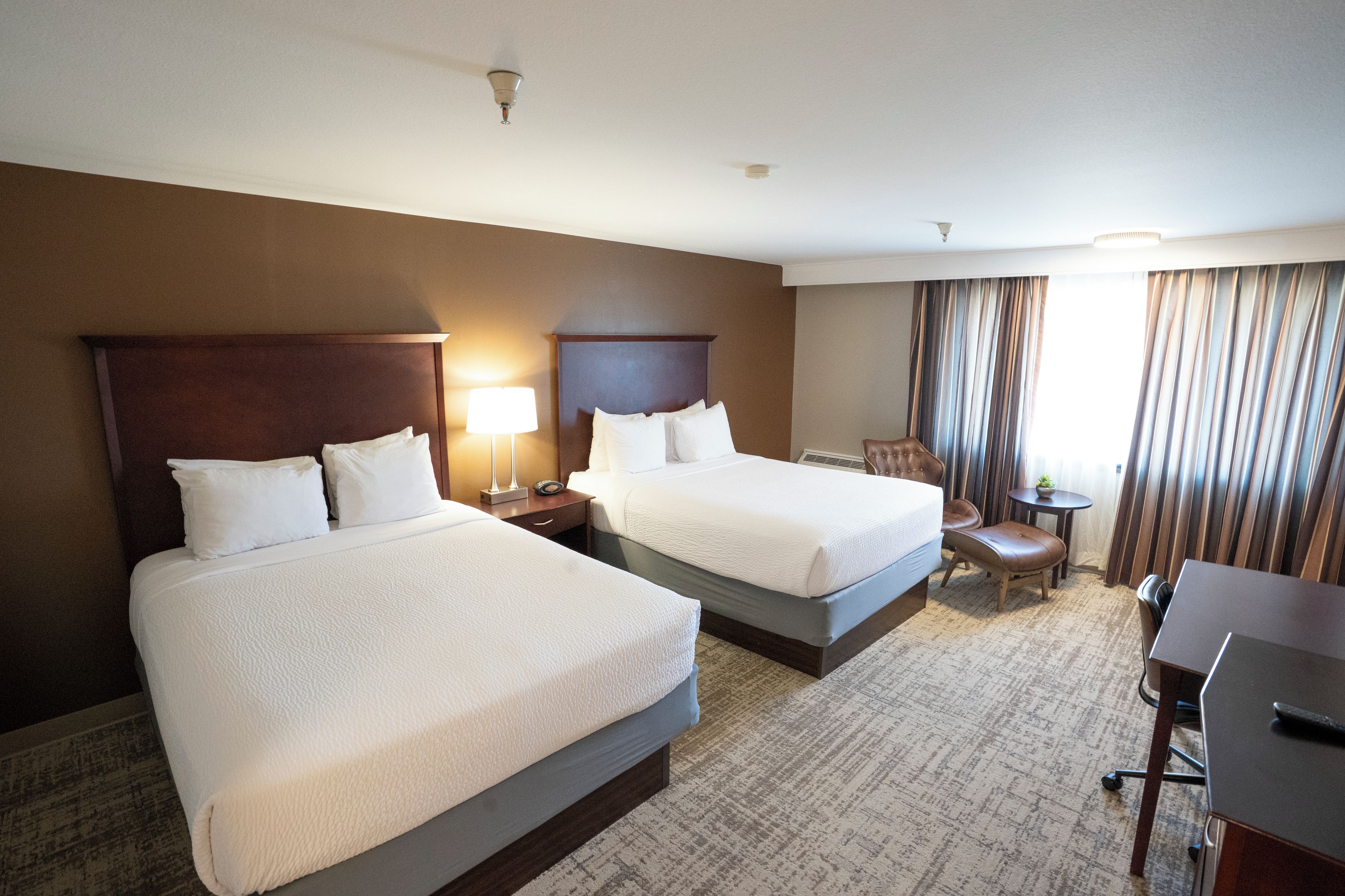 Habitación de hotel con dos camas, un escritorio, una silla, una lámpara y una ventana con cortinas.