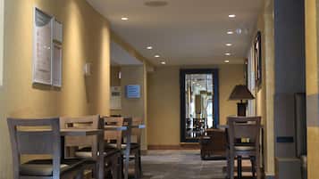 A hotel corridor with wooden chairs and tables, a lamp, and framed pictures on the walls.