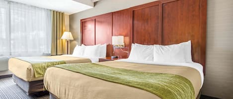 A hotel room with two beds, a large wooden headboard, a nightstand with a lamp, and a window with sheer curtains.