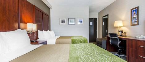 A hotel room with two beds, a desk, a chair, and a desk lamp. There are framed pictures on the wall and a door to the right.