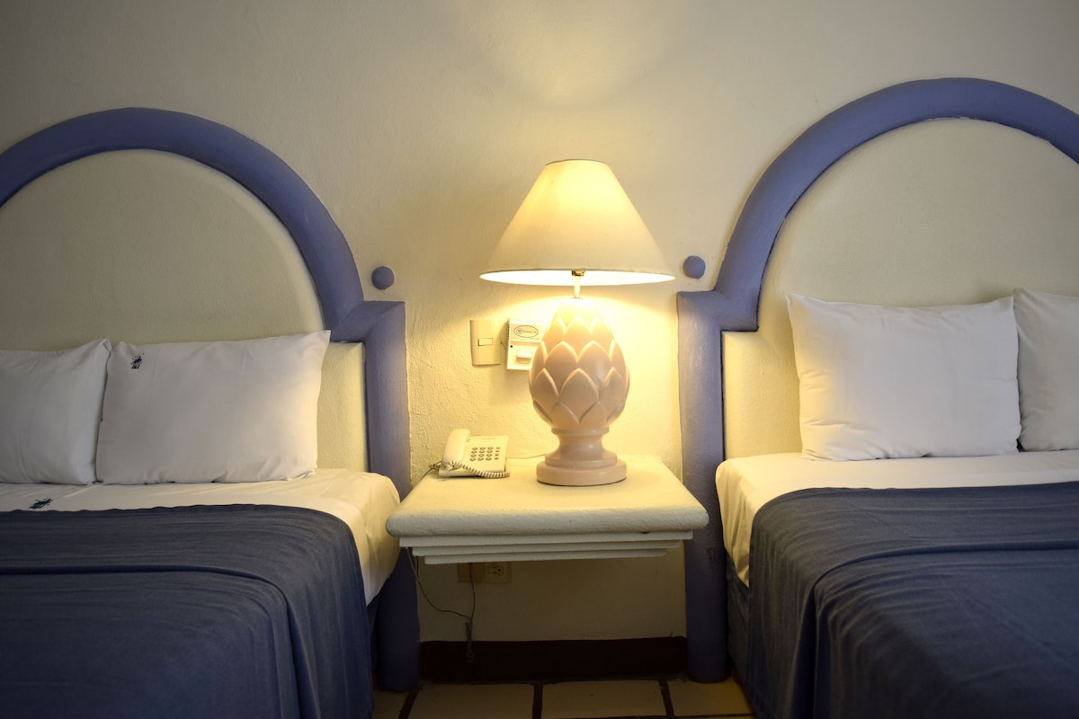 Quarto de hotel com duas camas, uma mesa de cabeceira com luminária e telefone.