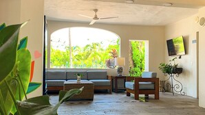 A spacious living area with a large window, a ceiling fan, a sofa, a coffee table, and a TV mounted on the wall. - Puerto de Luna Pet Friendly & Family Suites Hotel (Puerto Vallarta)