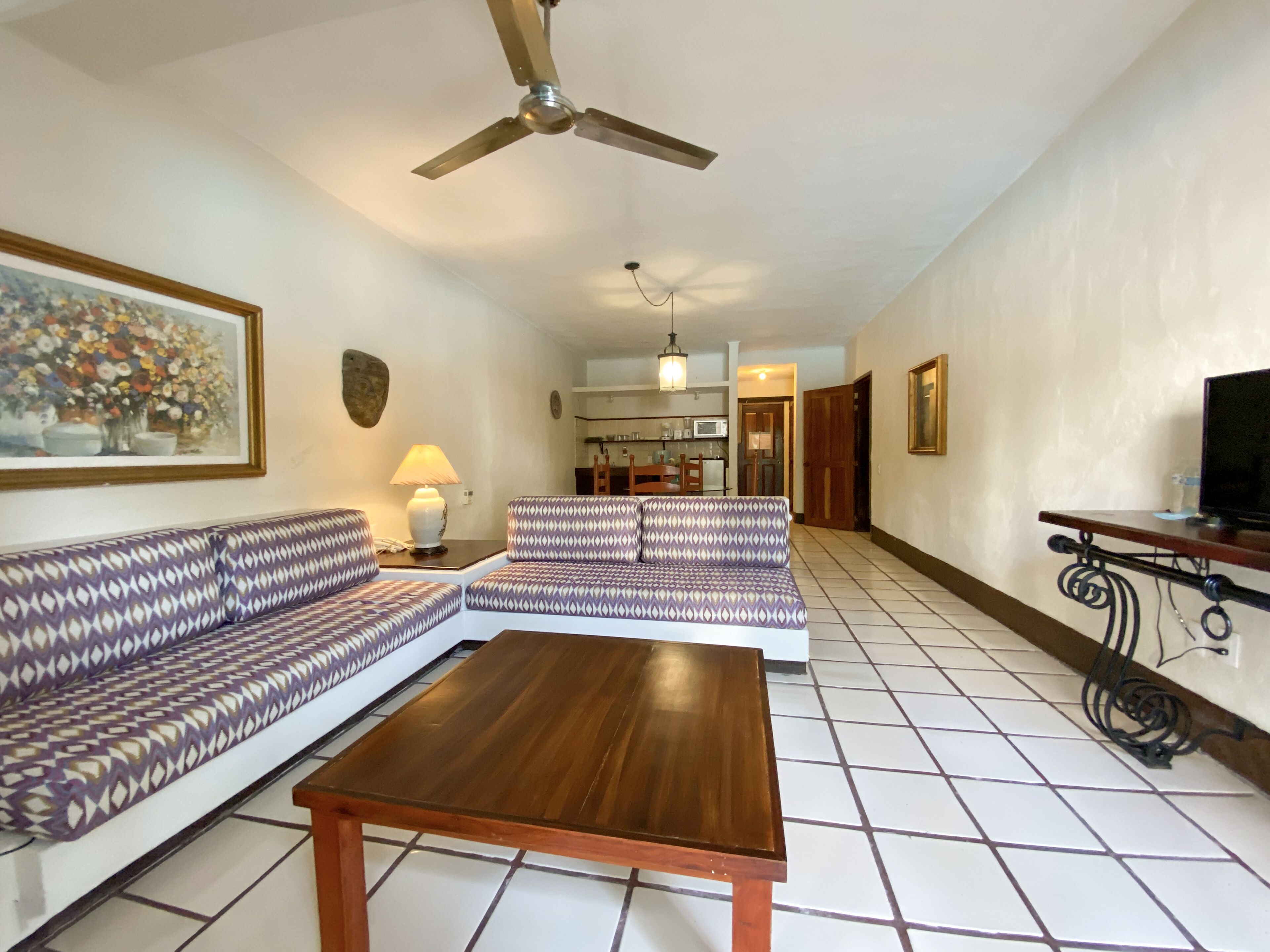 A living room with a sofa, a coffee table, a ceiling fan, a painting, and a kitchen in the background.