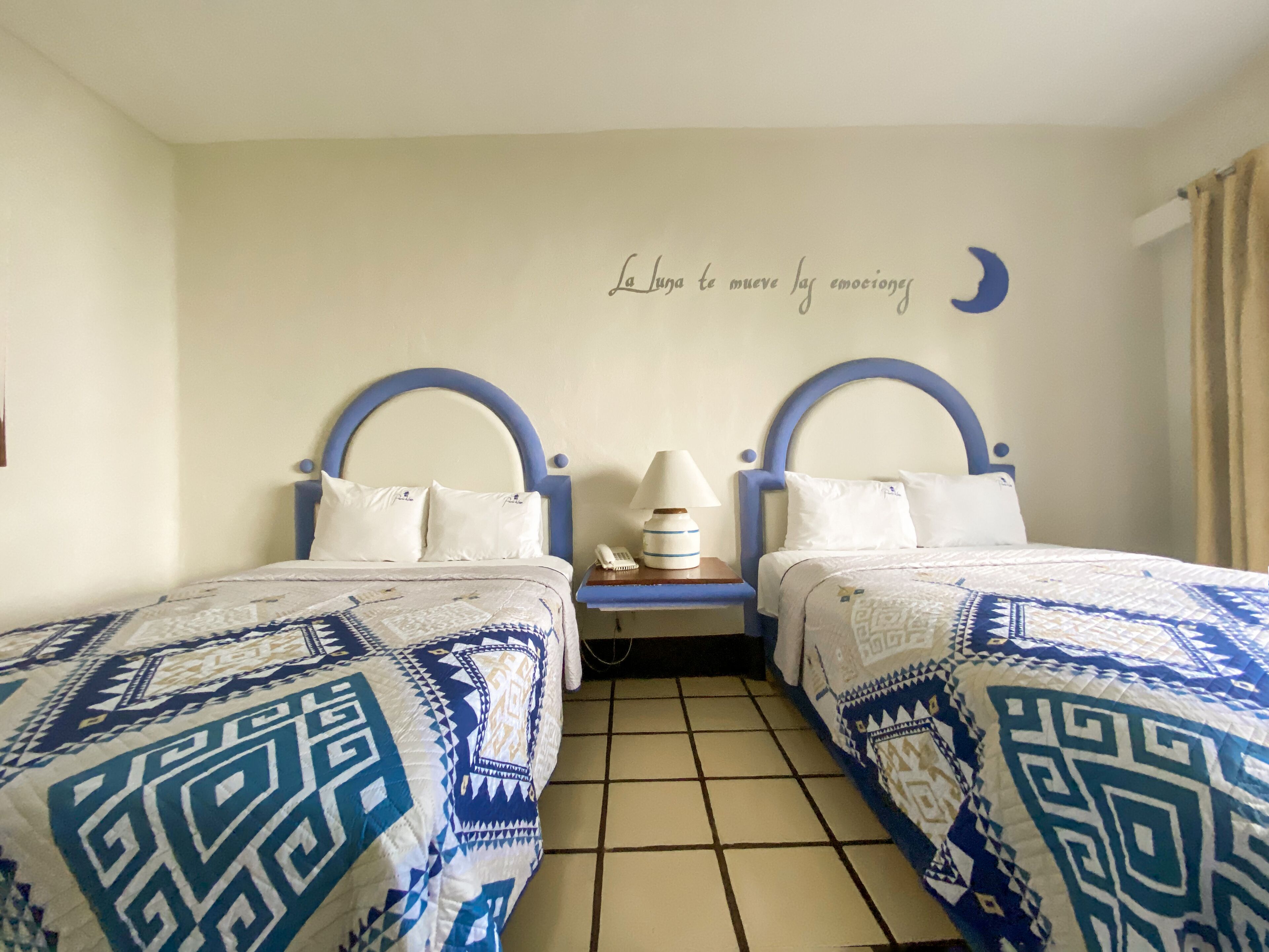 Een kamer met twee bedden, elk voorzien van een blauw hoofdeinde en witte beddengoed. Er is een wanddecoratie van een halve maan en een tekst die luidt: "La Luna te mueve la emoción".