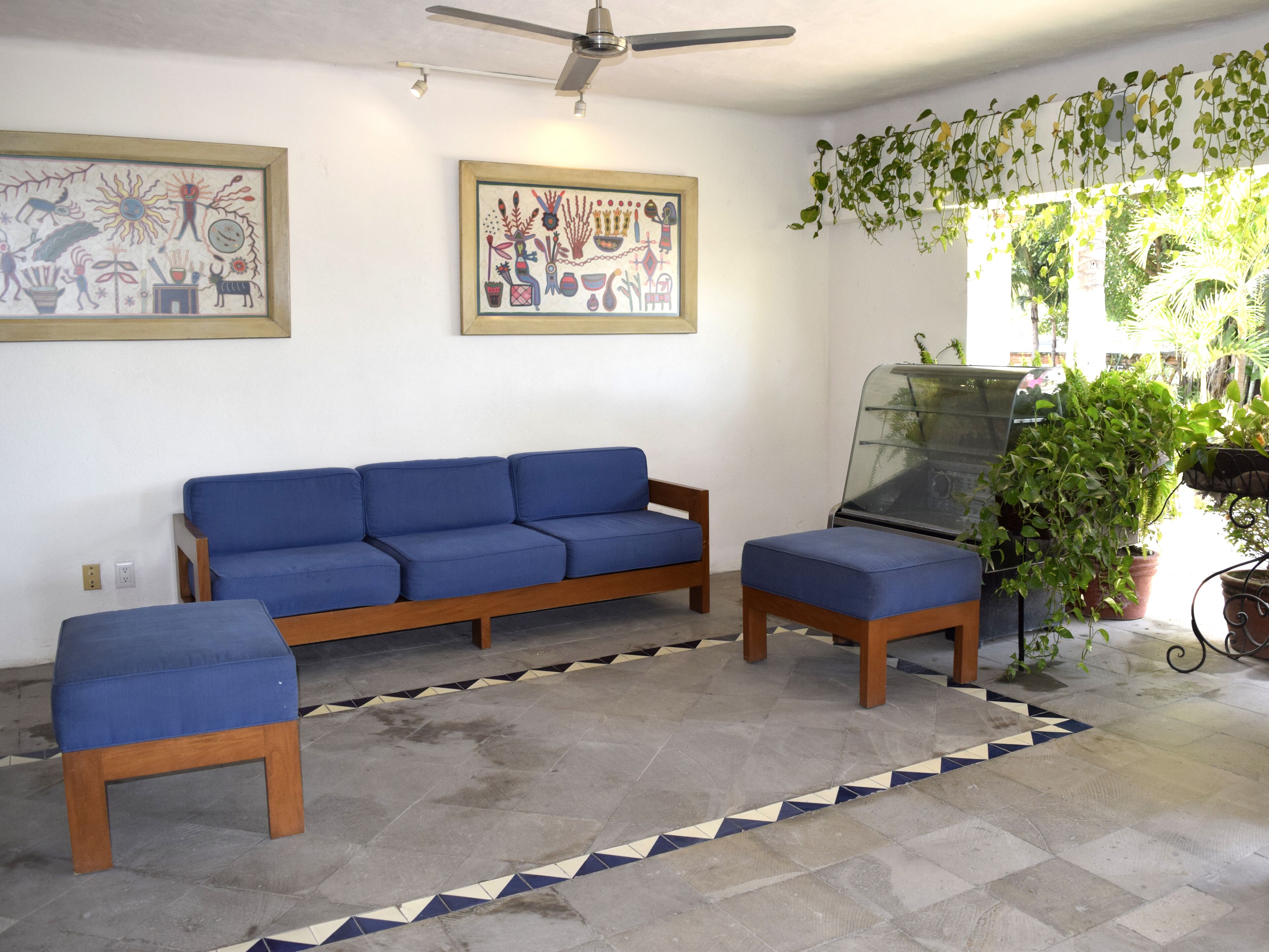 A living room with a blue sofa, matching ottoman, and a glass coffee table. There are two wall-mounted paintings and a ceiling fan.