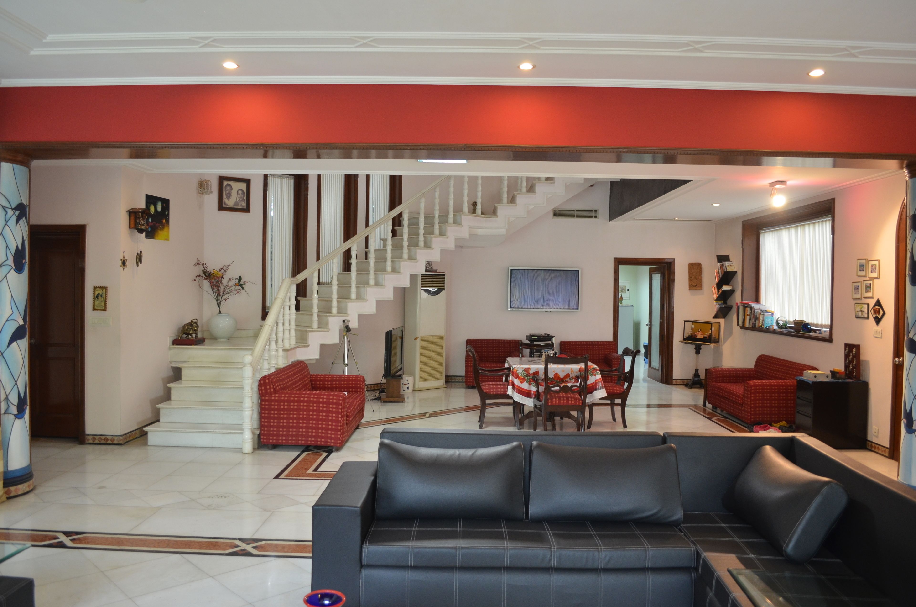 A spacious living room with a staircase, red and black sofas, a dining area, and a large window.
