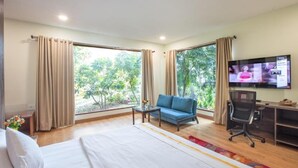 A hotel room with a bed, a blue sofa, a desk with a computer, a TV, and large windows with curtains. - The Radha Ashok (Mathura)