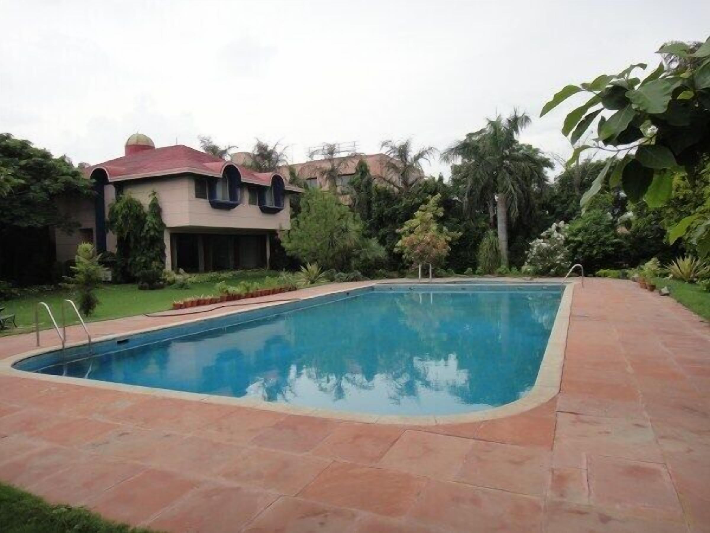 Una piscina rectangular con solárium de azulejos, rodeada de exuberante vegetación y un edificio con tejado rojo.