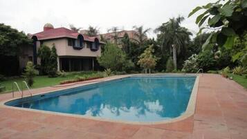 Una piscina rectangular con solárium de azulejos, rodeada de exuberante vegetación y un edificio con tejado rojo.