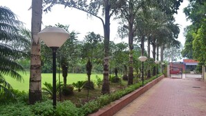 A paved pathway lined with trees and lamp posts, leading to a gated entrance. - The Radha Ashok (Mathura)