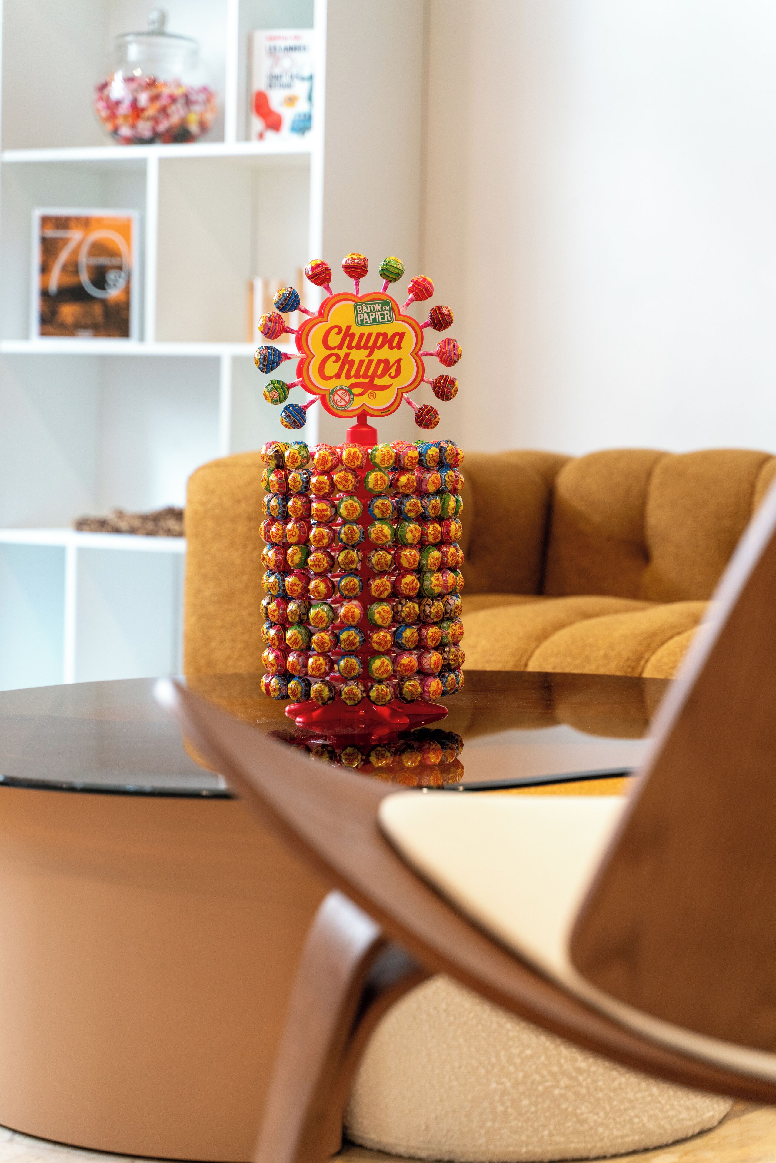 A decorative lollipop tree with a sign that reads "Chupa Chups" in a room with a brown sofa and white shelves in the background.