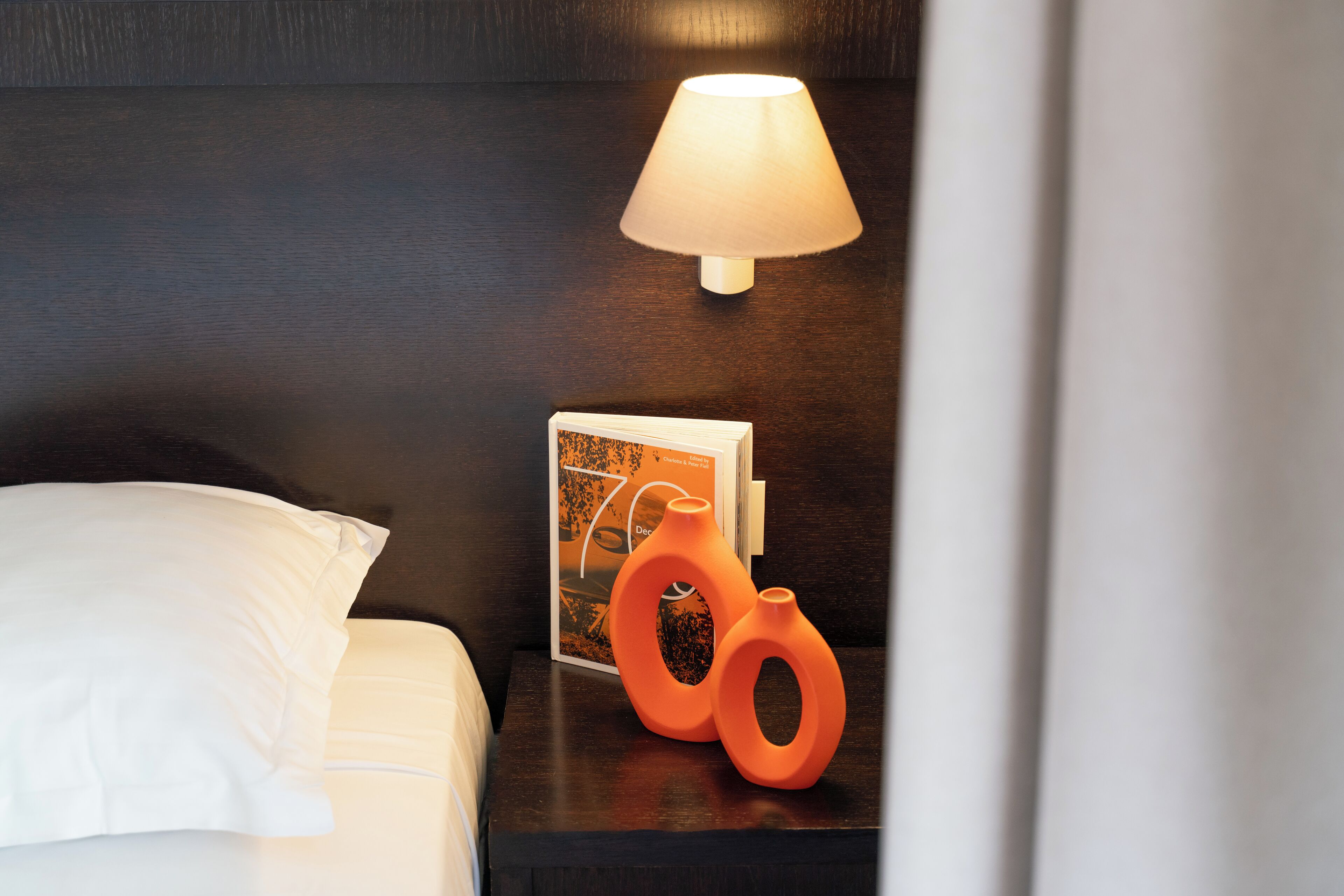 Una cama bien hecha con ropa blanca, un cabecero de madera oscura, una lámpara de noche y un pequeño florero naranja sobre una mesita de noche.