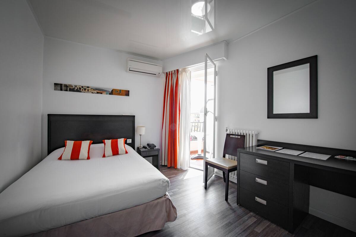 A modern hotel room with a large bed, a desk, and a chair. There is a window with curtains, a radiator, and a wall-mounted air conditioner.