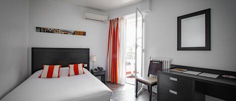 A modern hotel room with a large bed, a desk, and a chair. There is a window with curtains, a radiator, and a wall-mounted air conditioner.