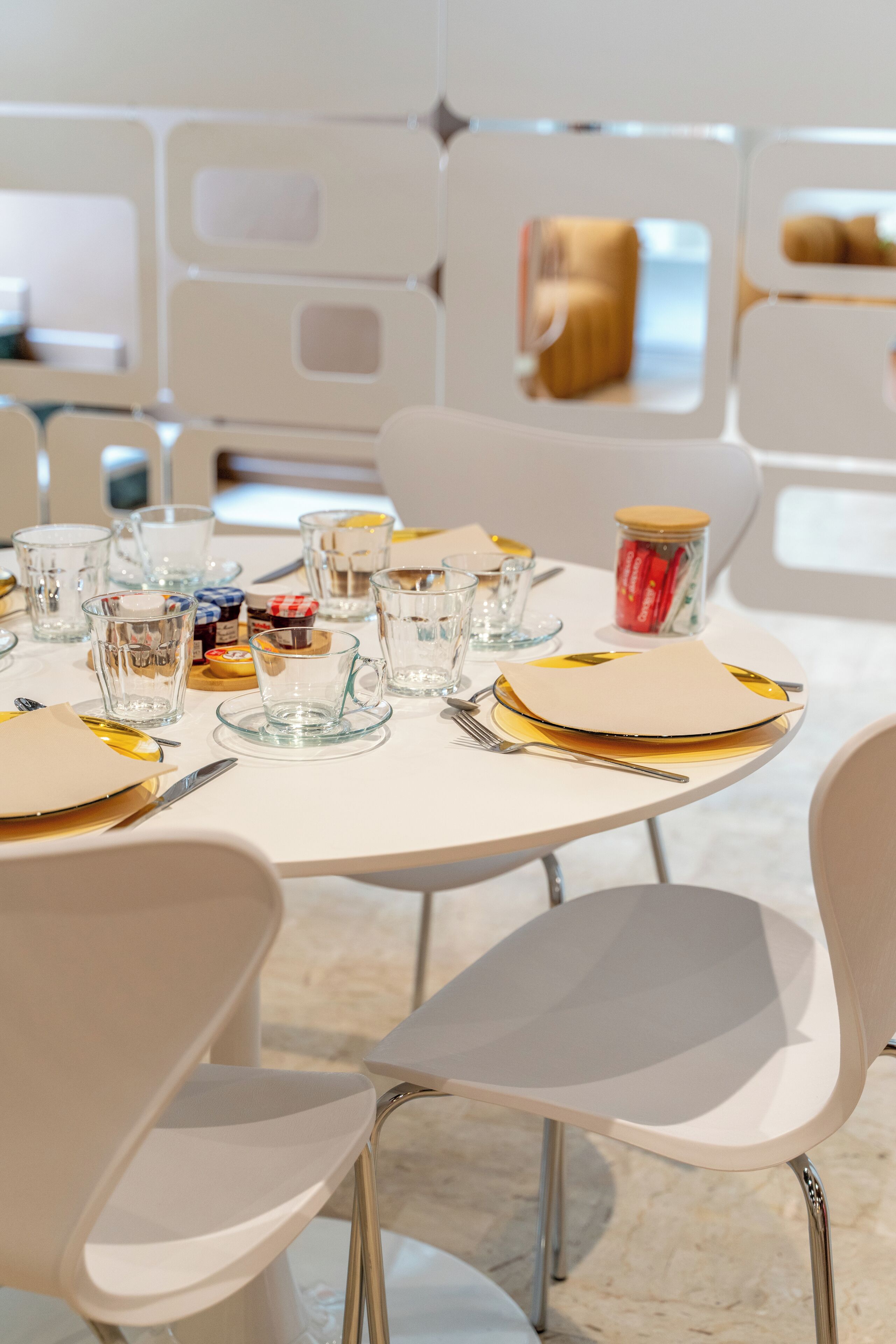 Una mesa de comedor dispuesta con vasos transparentes, platos y cubiertos, rodeada de sillas blancas.
