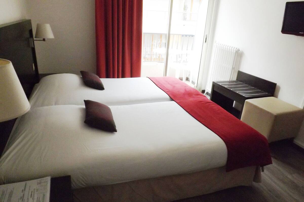 A hotel room with two beds, a red throw blanket, a bedside table, a lamp, and a view of the outside.