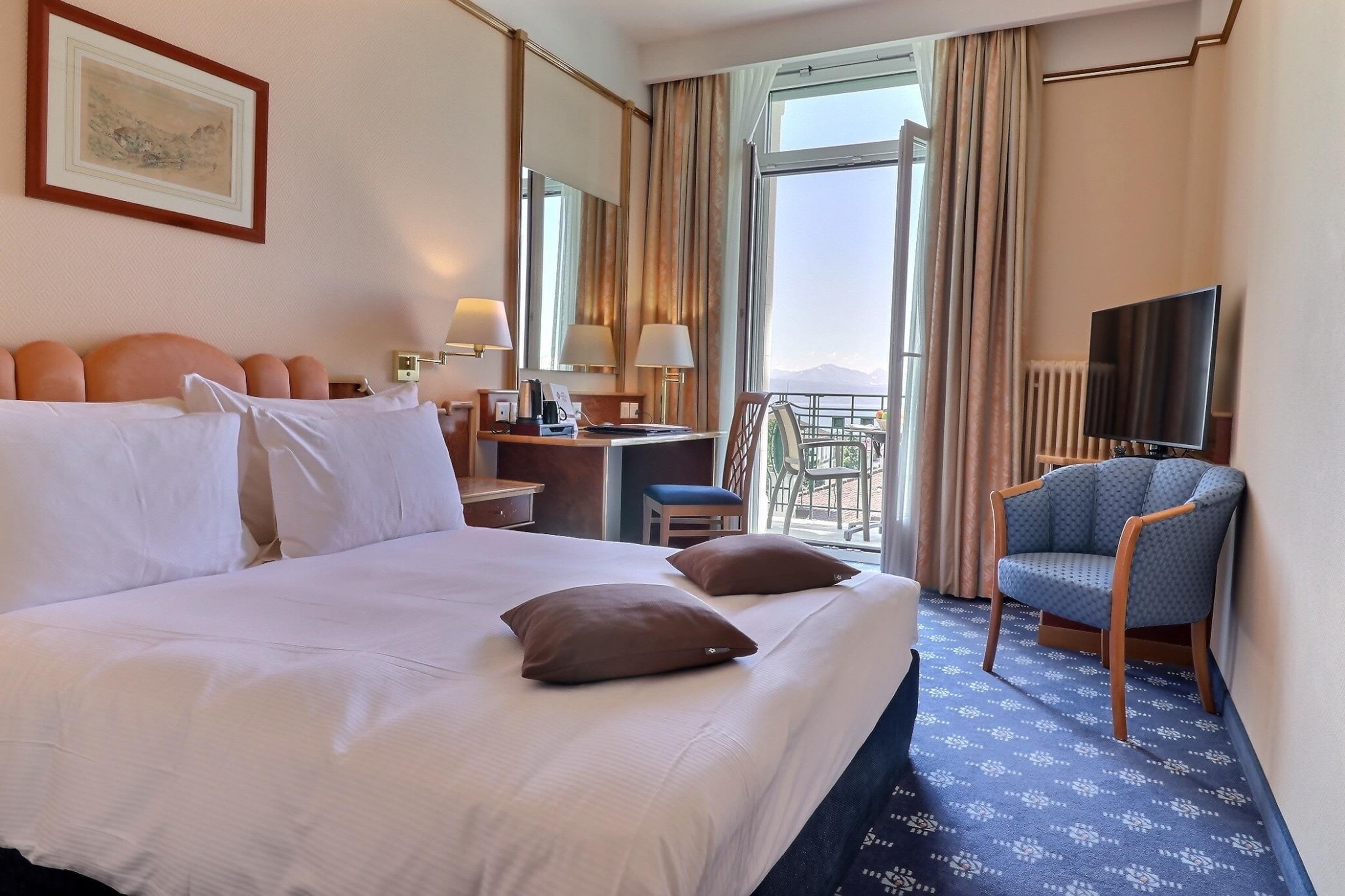 Ein Hotelzimmer mit einem großen Bett, einem Schreibtisch, einem Sessel, einem Fernseher und Blick ins Freie.