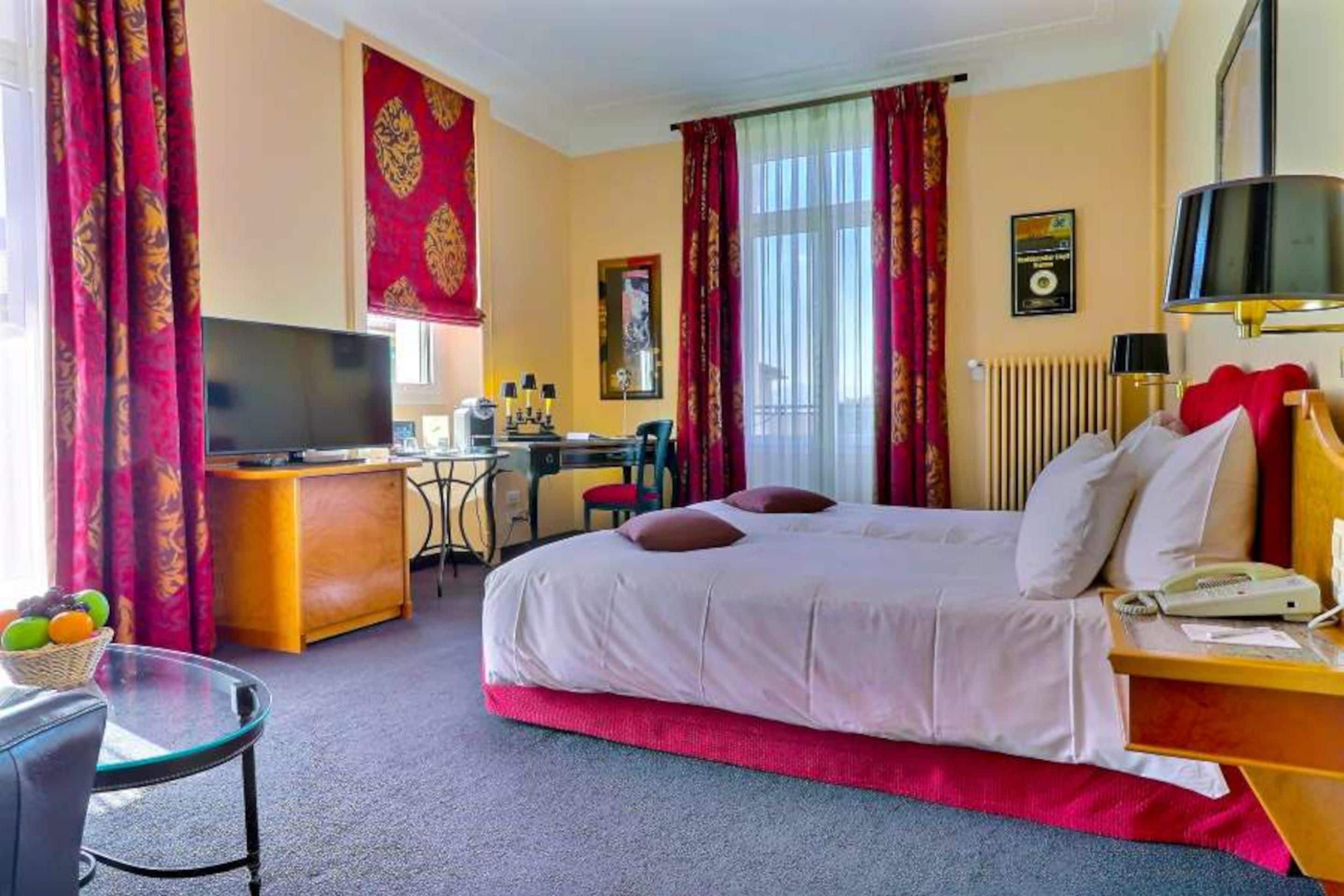 Ein Hotelzimmer mit Bett, Schreibtisch, Stuhl, Fernseher und einem Fenster mit Vorhängen.