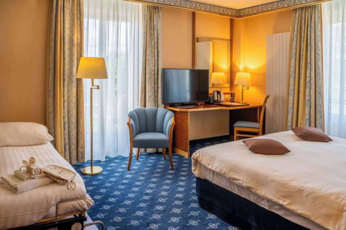 Une chambre d’hôtel avec un lit, un bureau avec une télévision, une chaise et un téléphone posé sur un petit chariot.