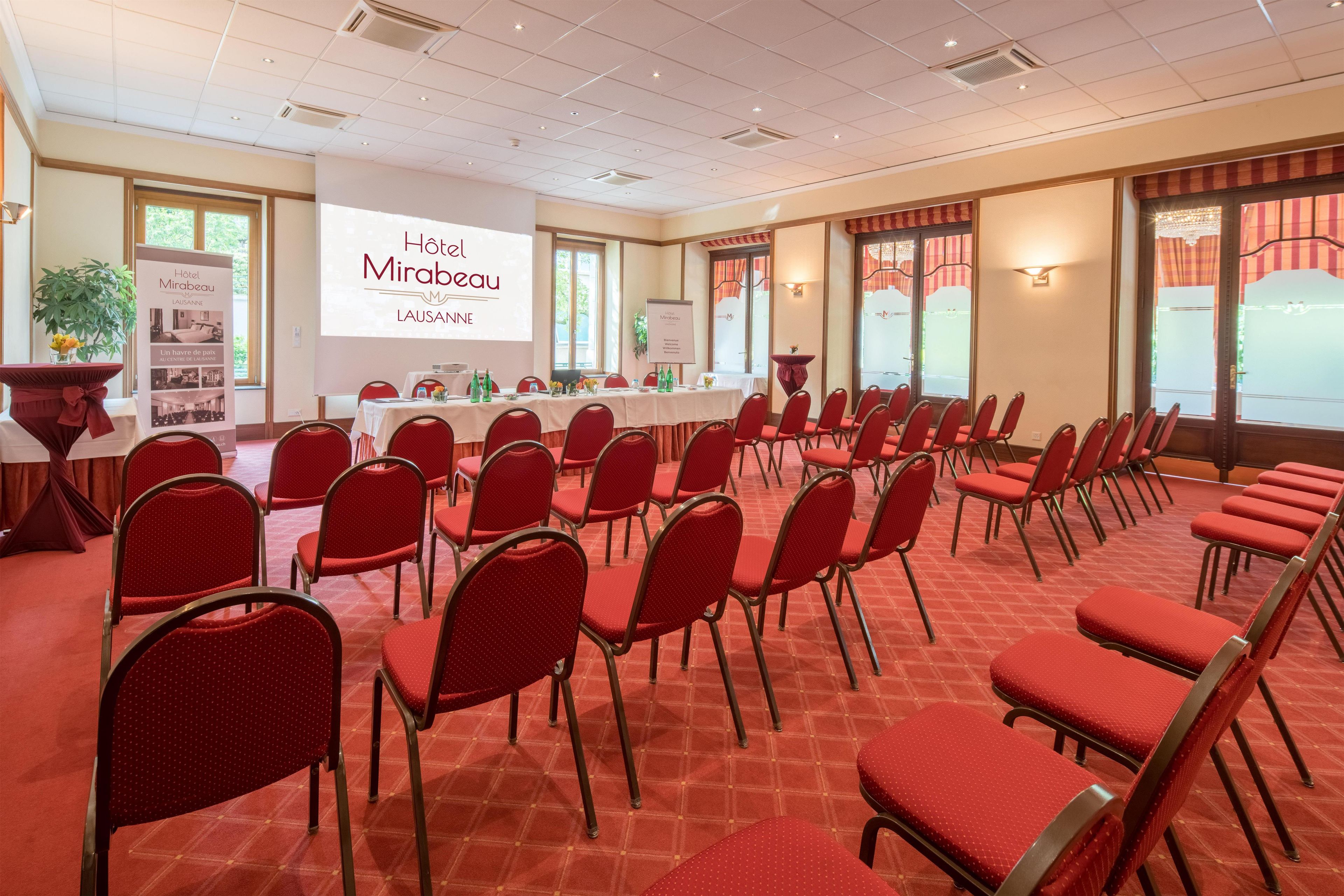 Une salle de conférence avec des rangées de chaises rouges, une table blanche sur laquelle est installé un écran de projection affichant « Hôtel Mirabeau Lausanne », et une grande fenêtre avec des rideaux.