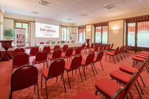 Un salón de conferencias con filas de sillas rojas, una mesa blanca con una pantalla de proyector mostrando 'Hotel Mirabeau Lausanne' y un ventanal con cortinas.