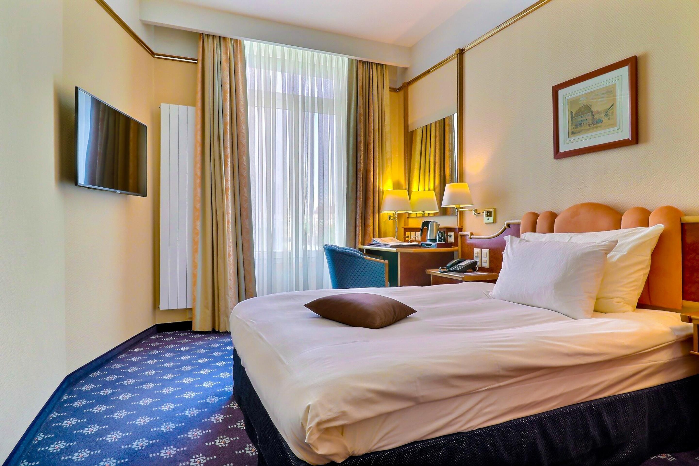 A hotel room with a bed, a desk with a lamp, a chair, a television, and a window with curtains.