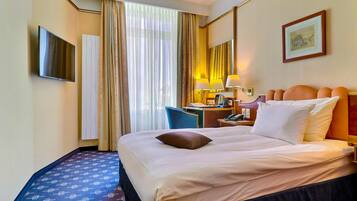 A hotel room with a bed, a desk with a lamp, a chair, a television, and a window with curtains.