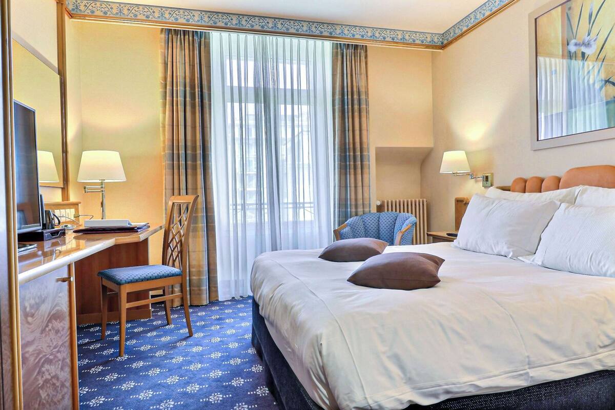 Une chambre d’hôtel avec un grand lit, un bureau avec une chaise, une lampe de bureau, une chaise et une fenêtre avec des rideaux.