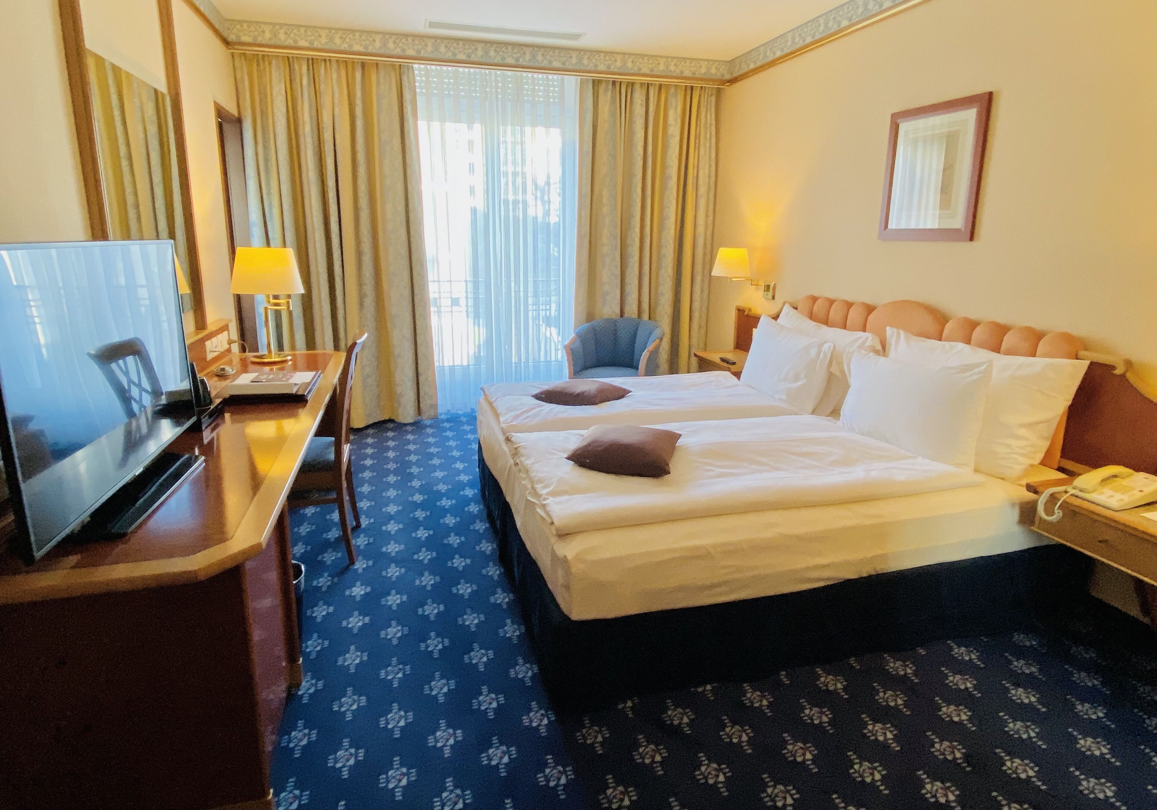 Une chambre d’hôtel avec un lit, un bureau avec une télévision, une chaise, une lampe et une fenêtre avec des rideaux.