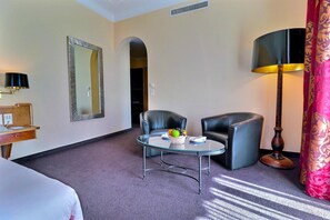 Una habitación de hotel con una cama, un escritorio con lámpara, una mesita de cristal con fruta, dos sillas, un espejo y una lámpara de pie.
