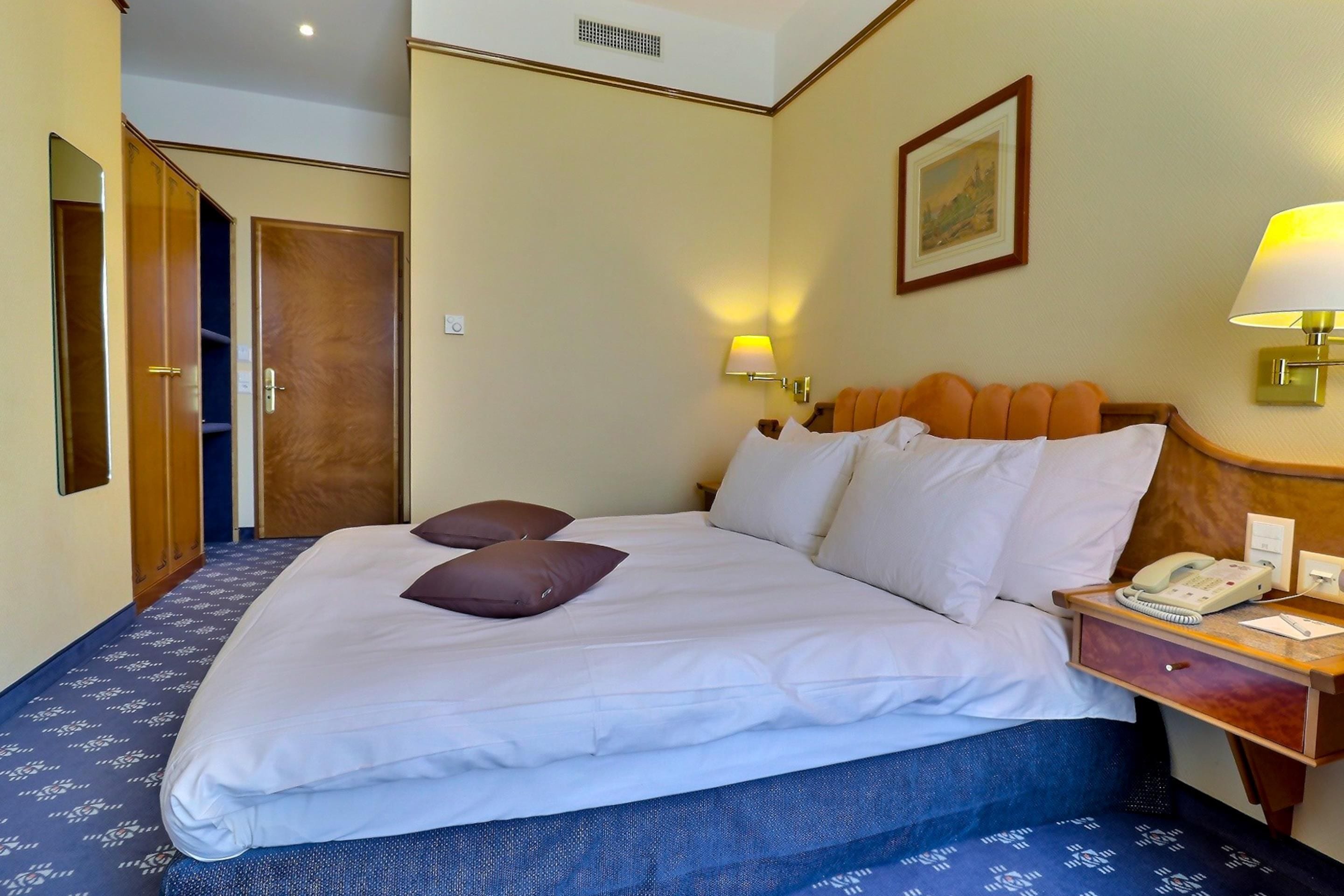A hotel room with a bed, pillows, a nightstand with a phone, and a painting on the wall.