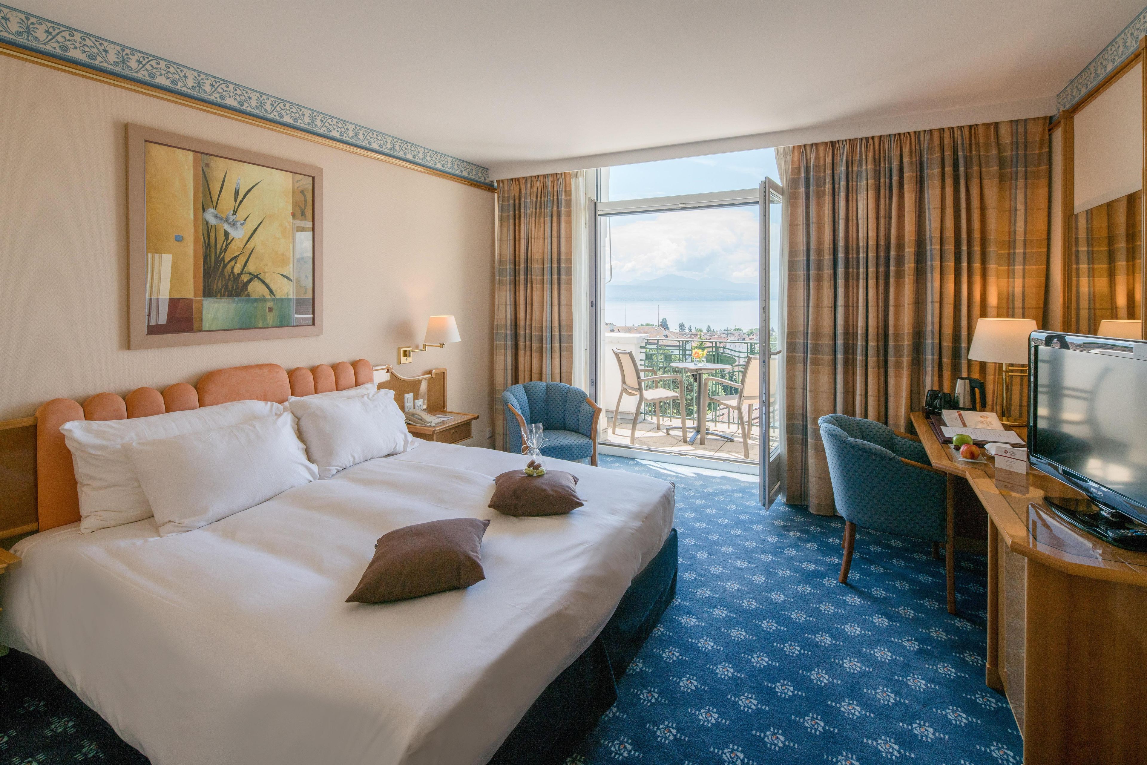 A hotel room with a large bed, a desk with a television, a chair, and a balcony with a view.