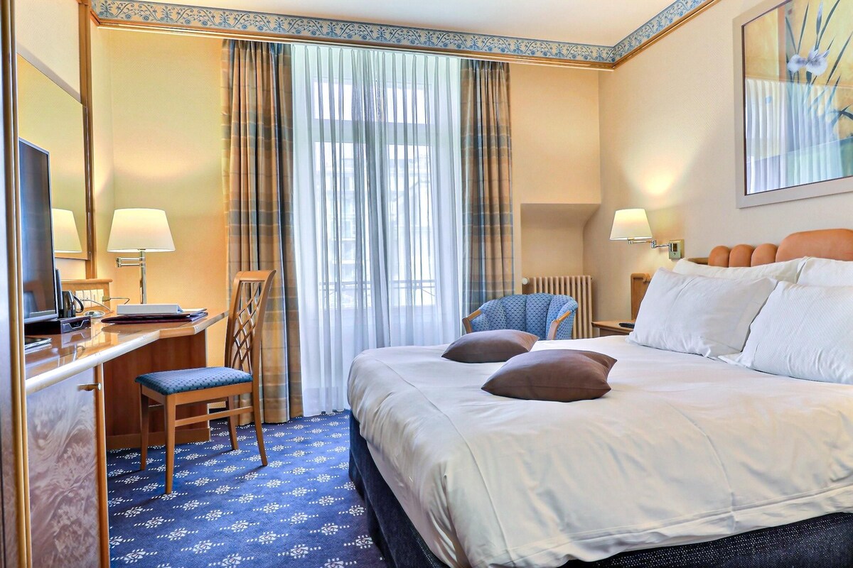 Habitación de hotel con cama, escritorio con computadora, silla, ventana con cortinas y lámpara de pared.