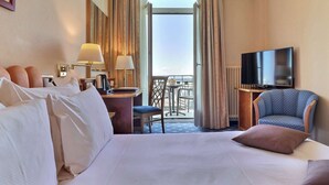 A hotel room with a large bed, a desk with a lamp, a television, and a chair. There is a balcony with a table and chairs visible through the open door.