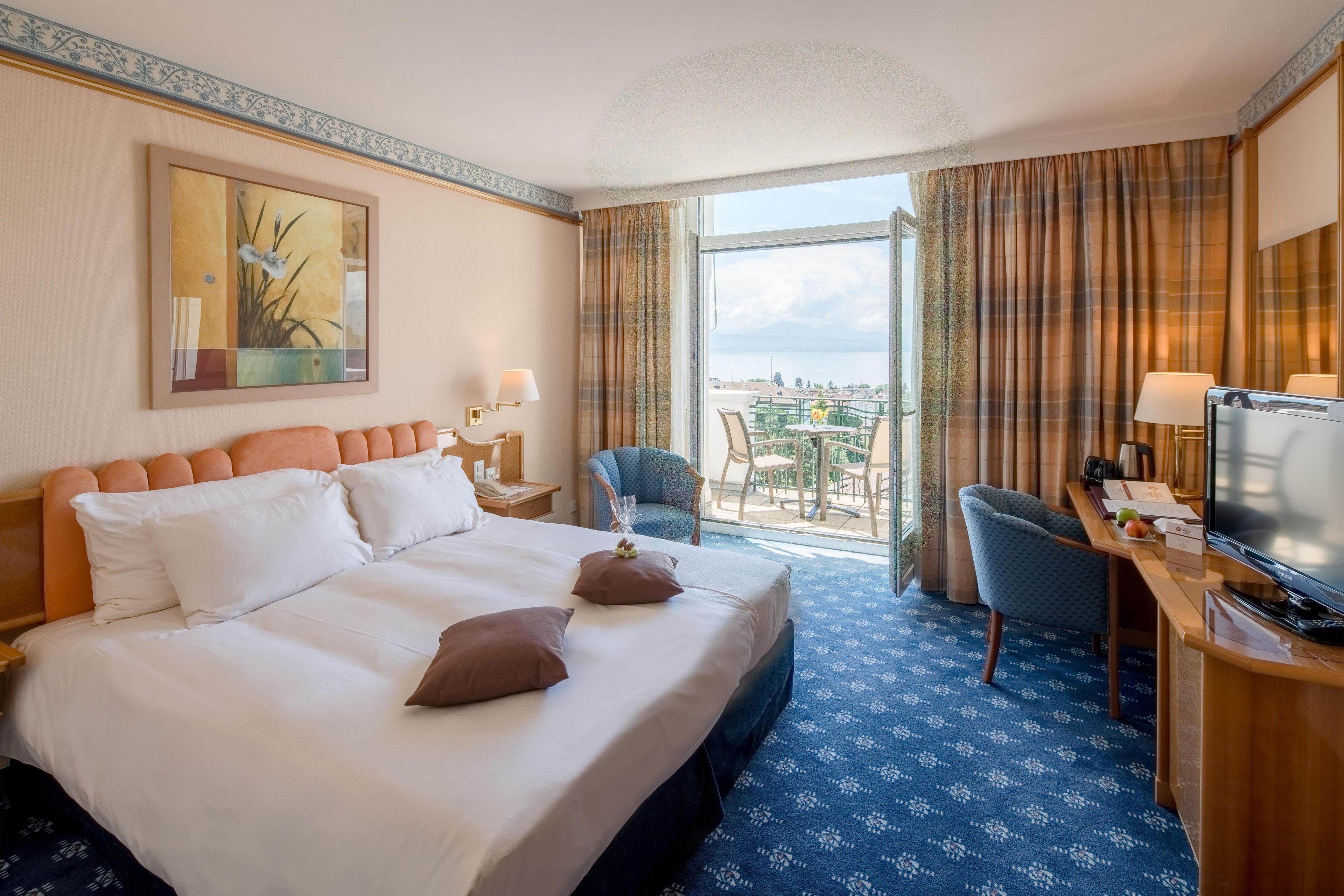 Habitación de hotel con una cama grande, un escritorio con televisor, una silla y vista a la ciudad por la ventana.