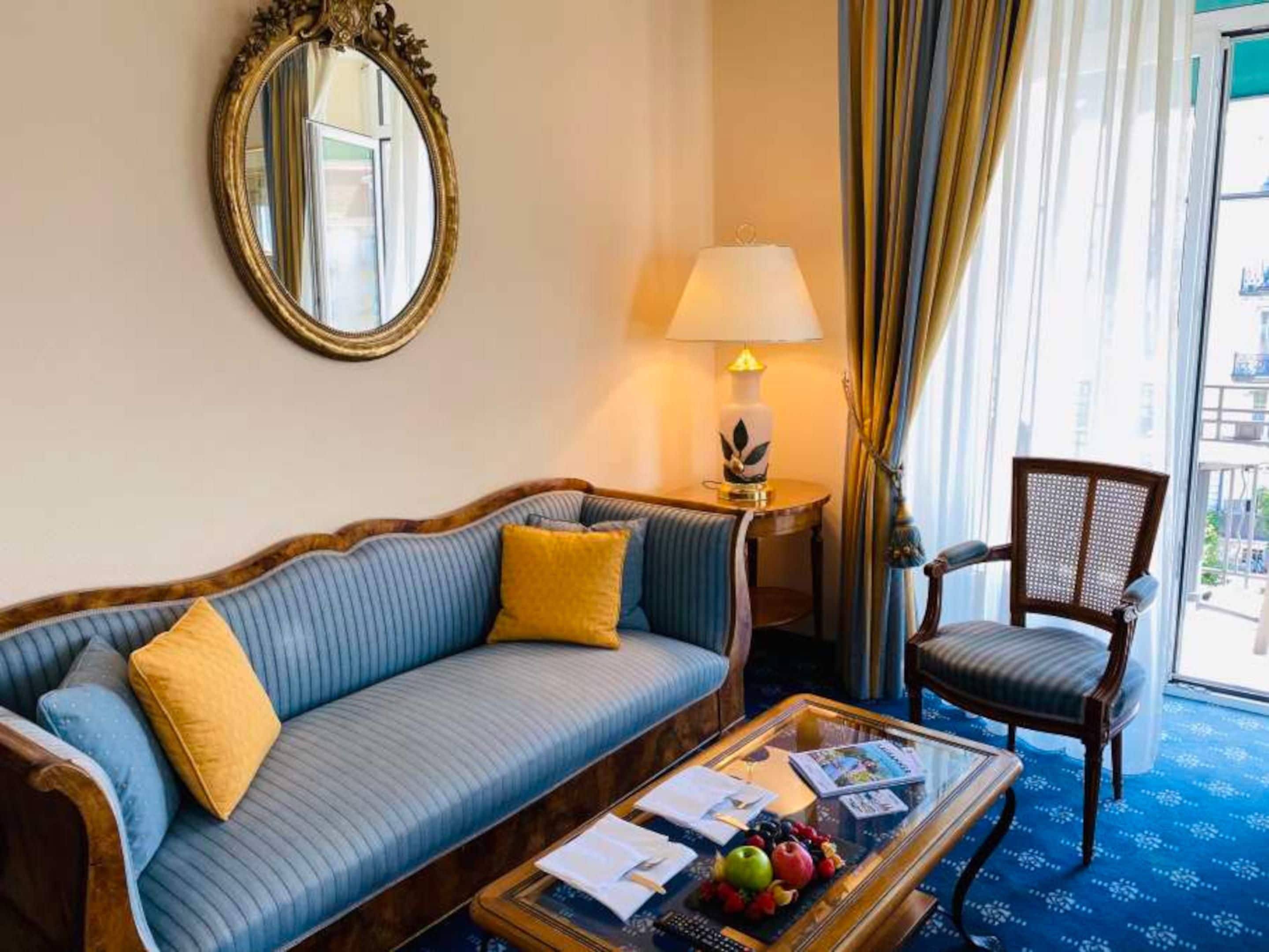 Une chambre d’hôtel avec un canapé, un fauteuil, une table basse avec des magazines et des fruits, une lampe et une fenêtre avec des rideaux.